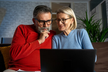 Happy mature couple using laptop at home. Happy couple with laptop and credit card sitting at home. Happy biracial couple sitting on sofa and using laptop in living room at home