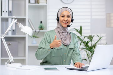 Smiling customer service representative wearing a headset gives a thumbs up while working at her office desk with a laptop ,and lamp in a modern workspace.