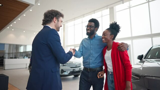 Caucasian salesman congratulating happy mixed-raced couple about successful purchase. Auto dealer giving car keys to satisfied woman. Pretty lady smiling with joy and hugging her husband.