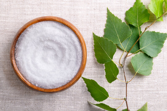 Birch sugar, table sweetener based on xylitol, in a wooden bowl on linen fabric. Also called xylite, a sugar substitute of natural origin with 40 percent fewer calories. A birch branch on the left.