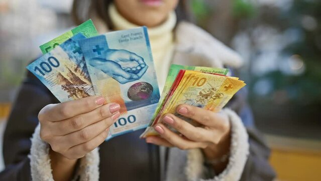 A young woman in outdoor attire holding swiss francs in front of a blurred natural backdrop.