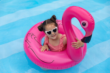 Happy child girl in pink swimsuit and sunglasses has fun in swimming ring flamingo in hotel swimming pool background. Travel, summer beach concept. Top view, copy space. Hotel spa resort advertisement