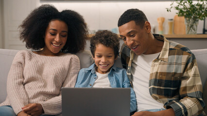 Happy African American family laughing woman mom female cheerful dad man male little boy son child kid watch movie cartoon using laptop computer pc playing game talking enjoy online shopping sofa home