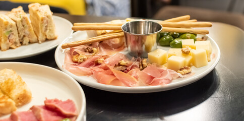 Appetizing snacks on white plate. Green olives, bruschetta, jamon, cheese, nuts close-up on blurred background of delicious snacks
