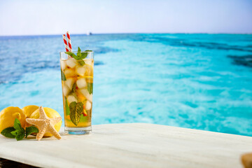 Glass of cold cocktail with lemon and mint on the wooden table with beach and sea and sky background.