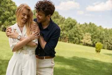 A beautiful young couple in elegant attire hugging tenderly in a green field, embodying love and serenity.