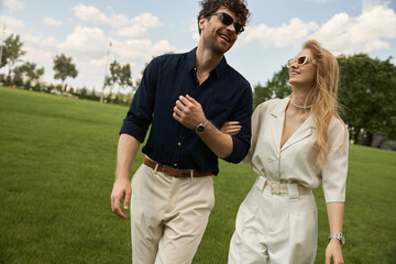 A beautiful young couple dressed in elegant clothing enjoys a leisurely walk in the park on a green...