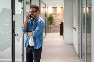 Positive male financial advisor with glasses making phone calls while standing in office corridor