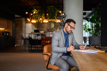 Serious male accountant with glasses examining spreadsheets and reports while sitting on chair at...