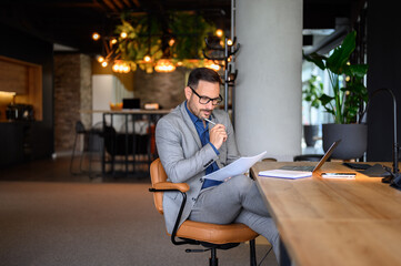 Focused male manager with notepad and laptop on desk reading document while working at desk in...
