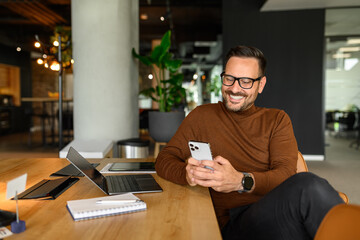 Young project manager reading e-mails over mobile phone while sitting by laptop and notepad at desk