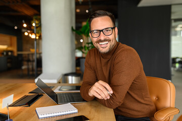 Male freelancer with laptop and notepad on desk smiling at camera while sitting on chair in office