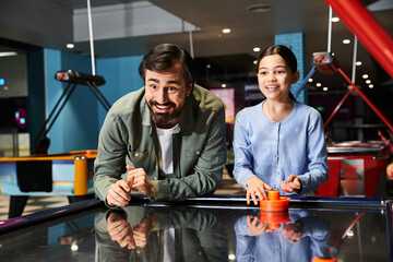 A dad and daughter engage in an intense game of air hockey at a gaming zone in a mall, having a fun...