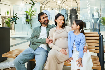 A pregnant woman and her daughter enjoy a moment together with man on a bench in a shopping mall during a weekend outing.