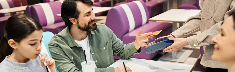A happy family sits at a restaurant table, happy man handing a credit card to a waitress