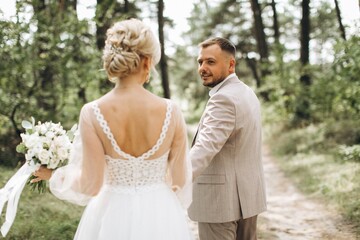 Fototapeta premium Wedding couple bride and groom walk hand in hand through a pine forest, sunlight filtering through the trees, highlighting their joyful expressions.