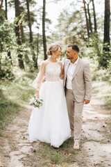 Wedding couple bride and groom walk hand in hand through a pine forest, sunlight filtering through the trees, highlighting their joyful expressions.