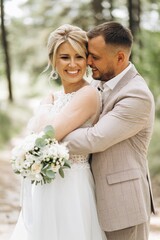 Wedding couple bride and groom hugging in pine forest, sunlight filtering through the trees, highlighting their joyful expressions.