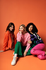 Three young multicultural women in stylish outfits sitting closely together on an orange background.