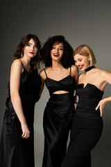 Three young women, each wearing a black dress, standing gracefully together against a neutral grey backdrop.