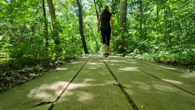 Fit middle adult woman is hiking in a lush green forest. She walks along a wooden plank path and is dressed casually in hiking gear. Located within Starved Rock State Park in LaSalle County, Illinois.