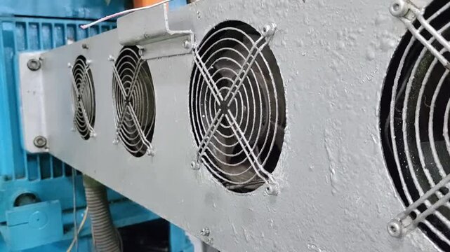 A close-up image of four industrial cooling fans mounted on a metal panel. The fans are circular and have a black metal grille. The panel is painted white and has a weathered appearance.