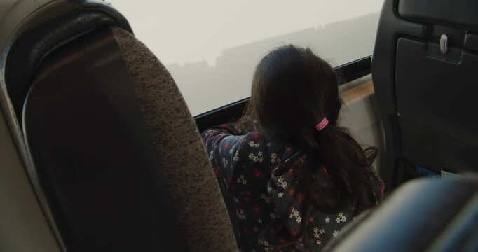 The girl sits on a soft seat, her small hands rest on the windowsill, as if she is trying to touch the world outside the window. View from the back of a child looking out the train window.