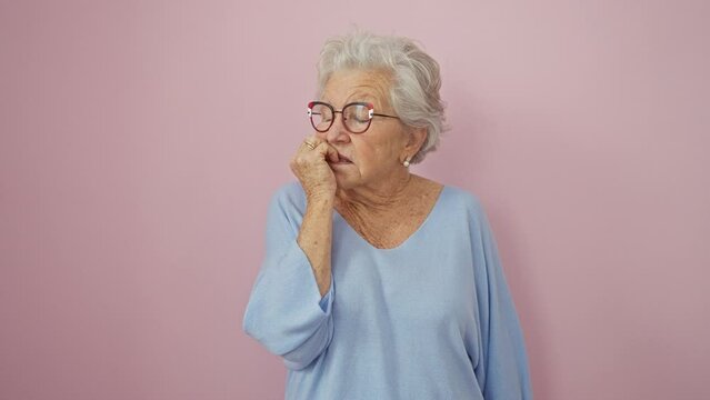 Grey-haired woman in glasses, biting nails and looking stressed over anxiety - fearful expression on pink isolated background