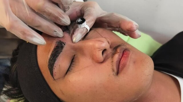 An esthetician using a microblade tool to make small cuts into the eyebrow of a customer. A Microblading technique to implant pigment. Male brow enhancement.