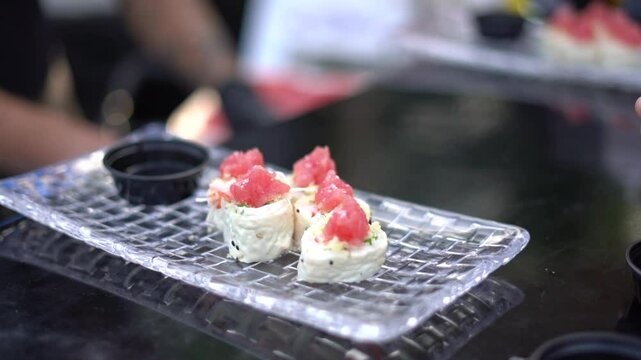 Un plato transparente con sushi de at&uacute;n fresco y en el fondo las manos de un chef preparando mas comida japonesa