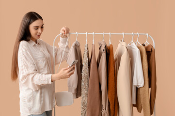 Happy female stylist with mobile phone and clothes on rack against beige background