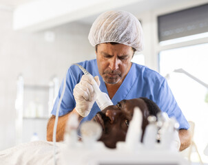 African man lying on couch during facial skin care procedure. Cosmetologist doctor using cold...