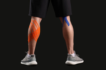 Young muscular man with physio tape on his legs against dark background, back view