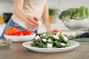 Young woman putting pieces of feta cheese in salad on kitchen