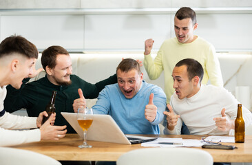 Fototapeta premium Excited friends gathered around laptop, celebrating successful investment made through online platform at casual home meeting with beer
