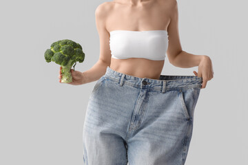 Beautiful young sporty woman in loose jeans with fresh broccoli on grey background, closeup. Weight...