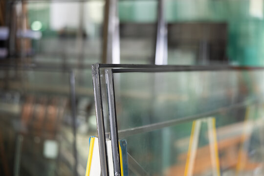Close-up of insulated glass units stacked on support stand in workshop, with various glass panels and industrial equipment in blurred background