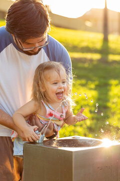 Young father lifting toddler up to drink at park water fountain bubbler