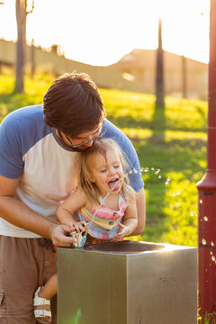 Young father lifting toddler up to drink at park water fountain bubbler