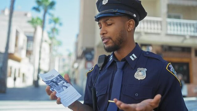 An african american police officer holding a missing person flyer on a sunny urban street.