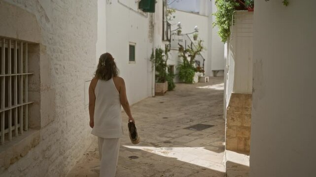 A young hispanic woman walking through the charming old town streets of locorotondo, puglia, italy, surrounded by white buildings and vibrant greenery.