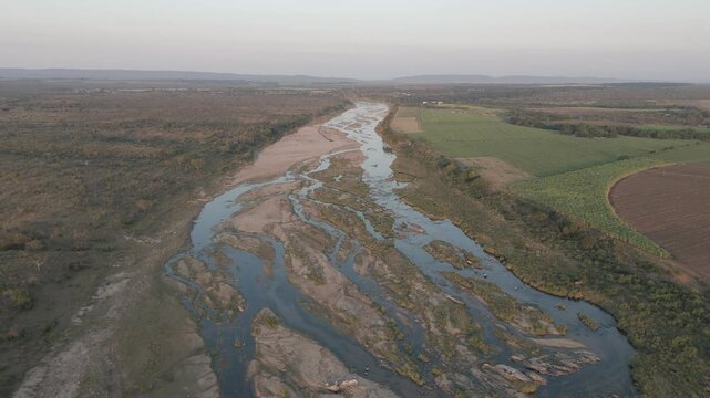Slow Drone rotating view reveals contrasting sugarcane farms, divided by a seasonal river, bordering Kruger National Park with scattered trees. South Africa.