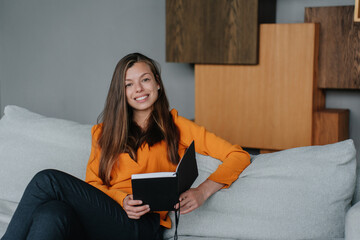 Smiling woman sitting on a couch, holding an open notebook, enjoying her time at home