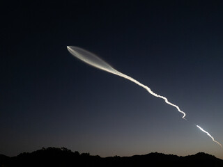 A bright white trail from a rocket launch stretches across the night sky as it ascends into space. The trail is visible against a dark blue sky, with a line of silhouetted hills.