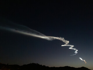 A bright white trail from a rocket launch stretches across the night sky as it ascends into space. The trail is visible against a dark blue sky, with a line of silhouetted hills.