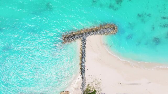 A static overhead shot capturing a T-shaped rock formation on a beach with clear blue water. The shot has calm waves gently hitting the rocks and the white sandy shore.