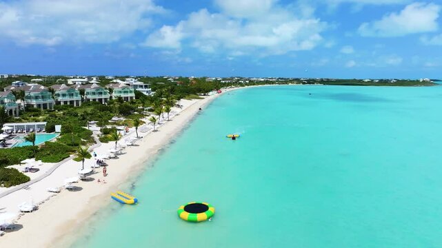 Reverse shot from the vibrant blue ocean with water tubing, transitioning onto the pristine white sandy beach with lilo and beachfront with luxurious villas and resorts, framed by green palm trees.