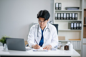 Confident young Asian male doctor in white medical uniform sit at desk working on computer. Smiling use laptop write