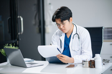 Confident young Asian male doctor in white medical uniform sit at desk working on computer. Smiling use laptop write