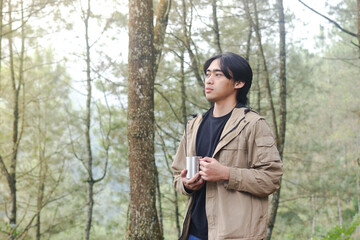 Portrait of excited Asian hiker man drinking water from stainless steel cup while trekking in nature forest